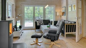 Een gezellig chaletinterieur met een houtkachel, een bank, een eettafel en een groot raam met uitzicht op het bos.