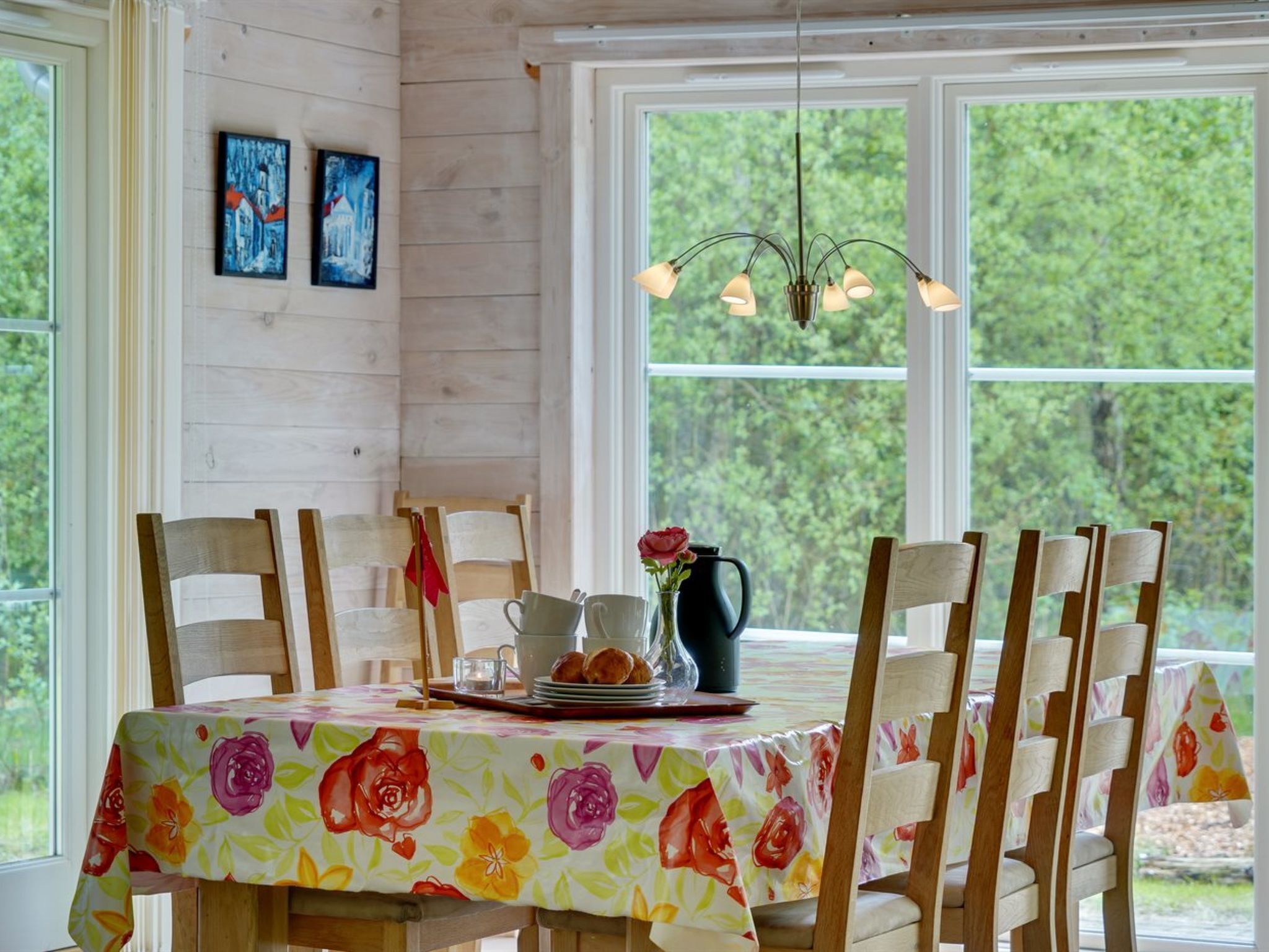 Un coin repas avec une table dressée pour quatre personnes, des chaises en bois, une nappe à motifs floraux, un lustre et des œuvres d’art accrochées au mur.