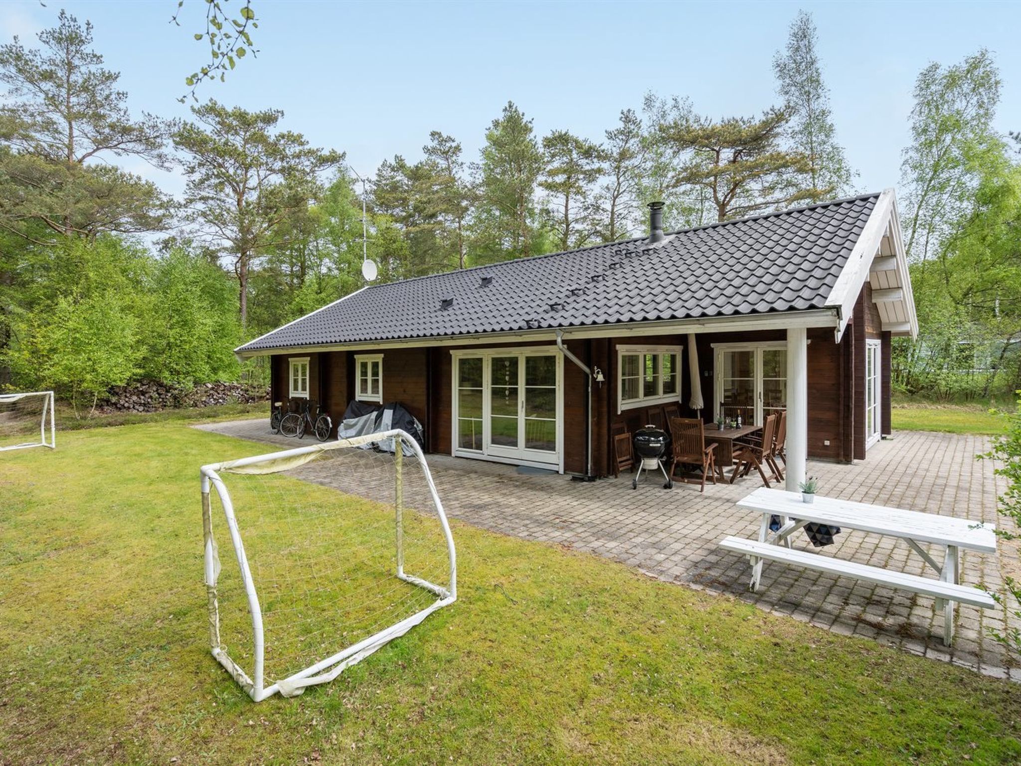 Une cabane en bois avec un toit en tuiles, une terrasse avec des meubles d’extérieur et un but de football dans la cour.
