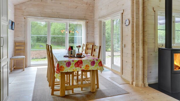 Een gezellig houten blokhutinterieur met een eettafel voor vier personen, een open haard en grote ramen met uitzicht op de omgeving.