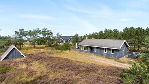 A modern house with a sloped roof, surrounded by a natural landscape with heather and trees.
