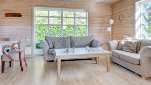 Et hyggeligt værelse med træindretning, et stort vindue med udsigt til grønne områder, en grå sofa, et træbord og en stol med hvid pude.