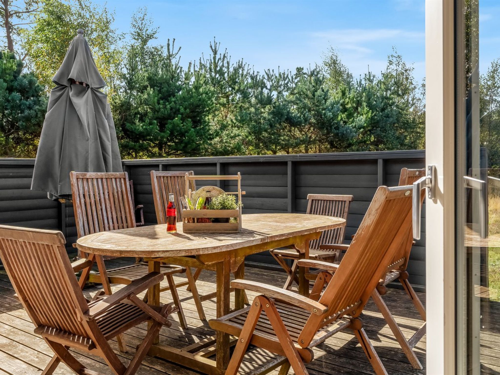 Une terrasse en bois avec une table ronde et six chaises, une bouteille de vin rouge et un panier rempli de plantes vertes.