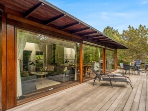 A modern outdoor deck with a pergola, glass walls, and a dining area with chairs and a table.