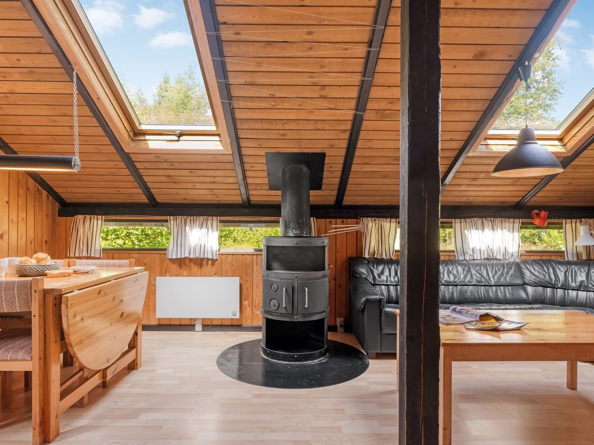 A cozy cabin with a central wood-burning stove, wooden furniture, and large windows.