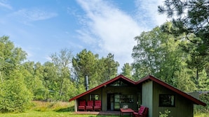 A cabin with a red roof, a porch with red chairs, and a grassy yard surrounded by trees.