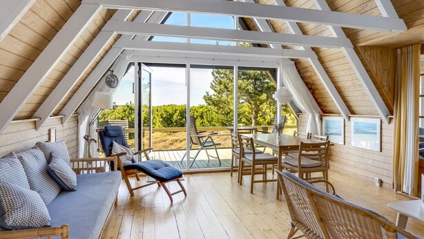 Ein Schlafzimmer im Dachgeschoss mit Holzboden, einem Sofa, einem Esstisch und einem großen Fenster mit Blick auf einen Garten.