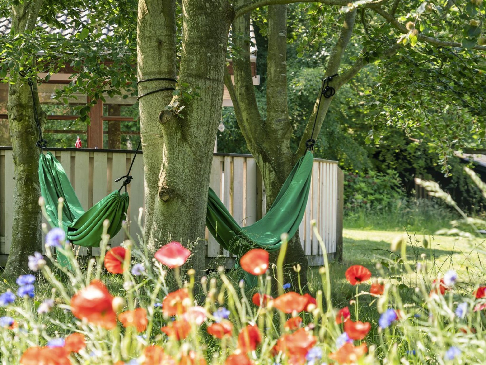 Een hangmat gespannen tussen twee bomen in een tuin met een houten omheining en bloeiende bloemen.