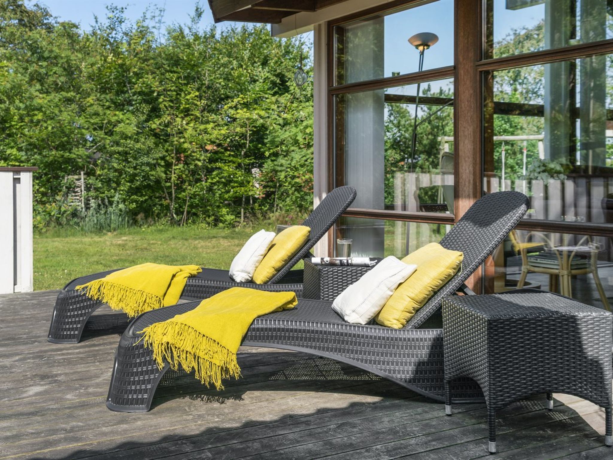 A sun lounger with a yellow throw blanket and pillows, positioned on a wooden deck with a glass wall and a view of a garden.