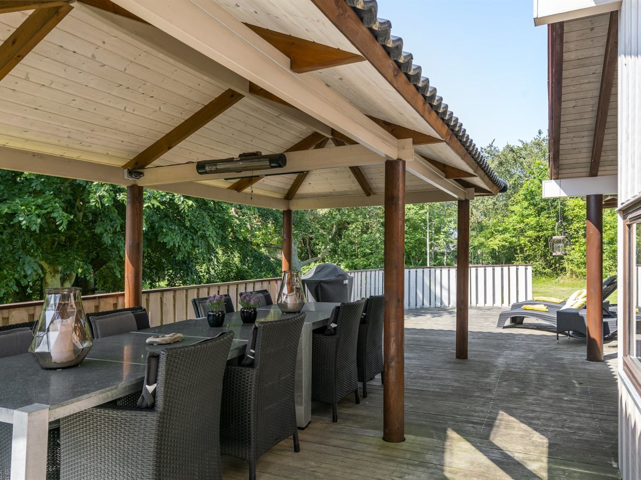 Een overdekt terras buiten met een tafel, stoelen, een barbecue en uitzicht op de omliggende begroeiing.