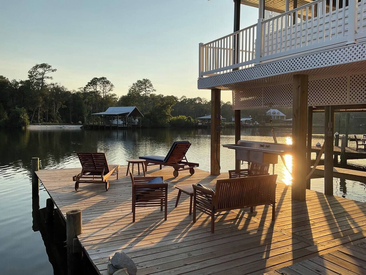 Una terraza de madera con sillones de descanso y una mesa, con vista a una masa de agua tranquila que incluye un muelle y edificios al fondo.