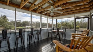 Une terrasse extérieure couverte, dotée de chaises et de tables en bois, et de grandes fenêtres offrant une vue sur le paysage environnant.