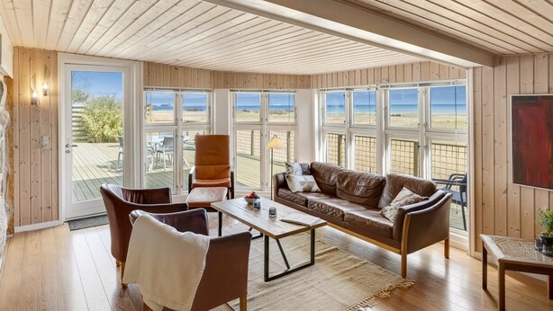 Una habitación amplia con techo alto, paredes de madera, un sofá de cuero, una mesa de comedor y grandes ventanales con vista a la playa.