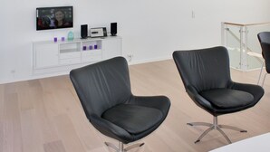 Un espace de bureau moderne avec deux fauteuils en cuir noir, une console blanche équipée d’enceintes et d’un téléviseur, et une rambarde en verre près de la fenêtre.