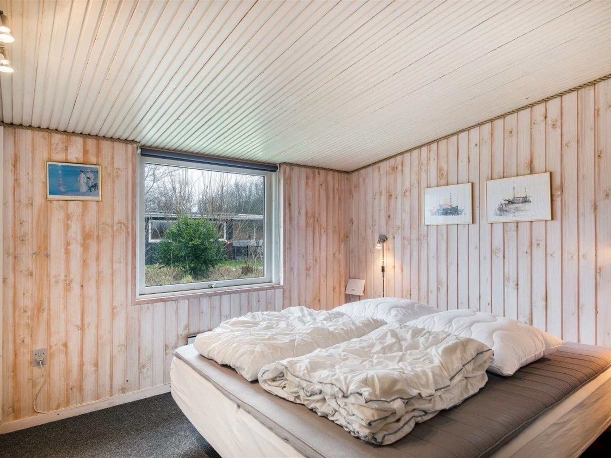 A bedroom with wooden walls, a bed with white bedding, a window with a view of greenery, and two wall-mounted lamps.