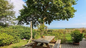 A wooden picnic table with benches, surrounded by lush greenery and a view of the sea.