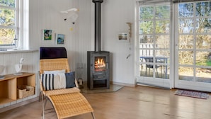 Une pièce avec un poêle à bois, un fauteuil en osier, une fenêtre et une porte donnant sur l’extérieur.