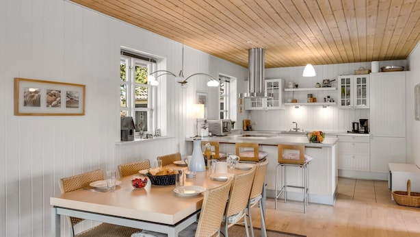 Une cuisine moderne avec des meubles blancs, un plafond en bois et un coin repas comprenant des chaises et une table en osier.