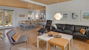 Un vaste espace de vie doté d’un plafond en bois, d’un canapé en cuir noir, d’une table à manger et d’une cuisine à l’arrière-plan.