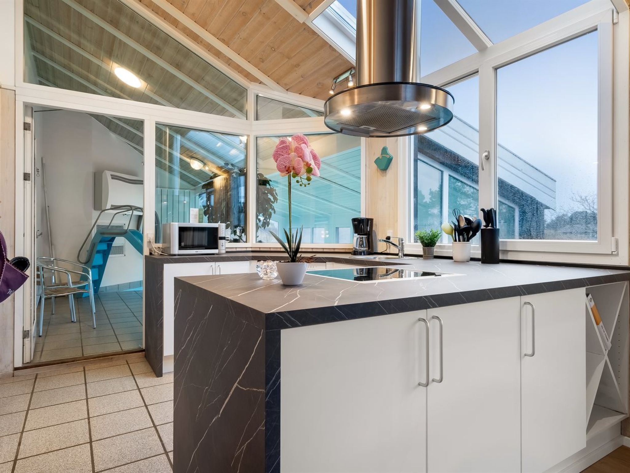 Een moderne keuken met een groot eiland, een roestvrijstalen afzuigkap en uitzicht op buiten door grote ramen.