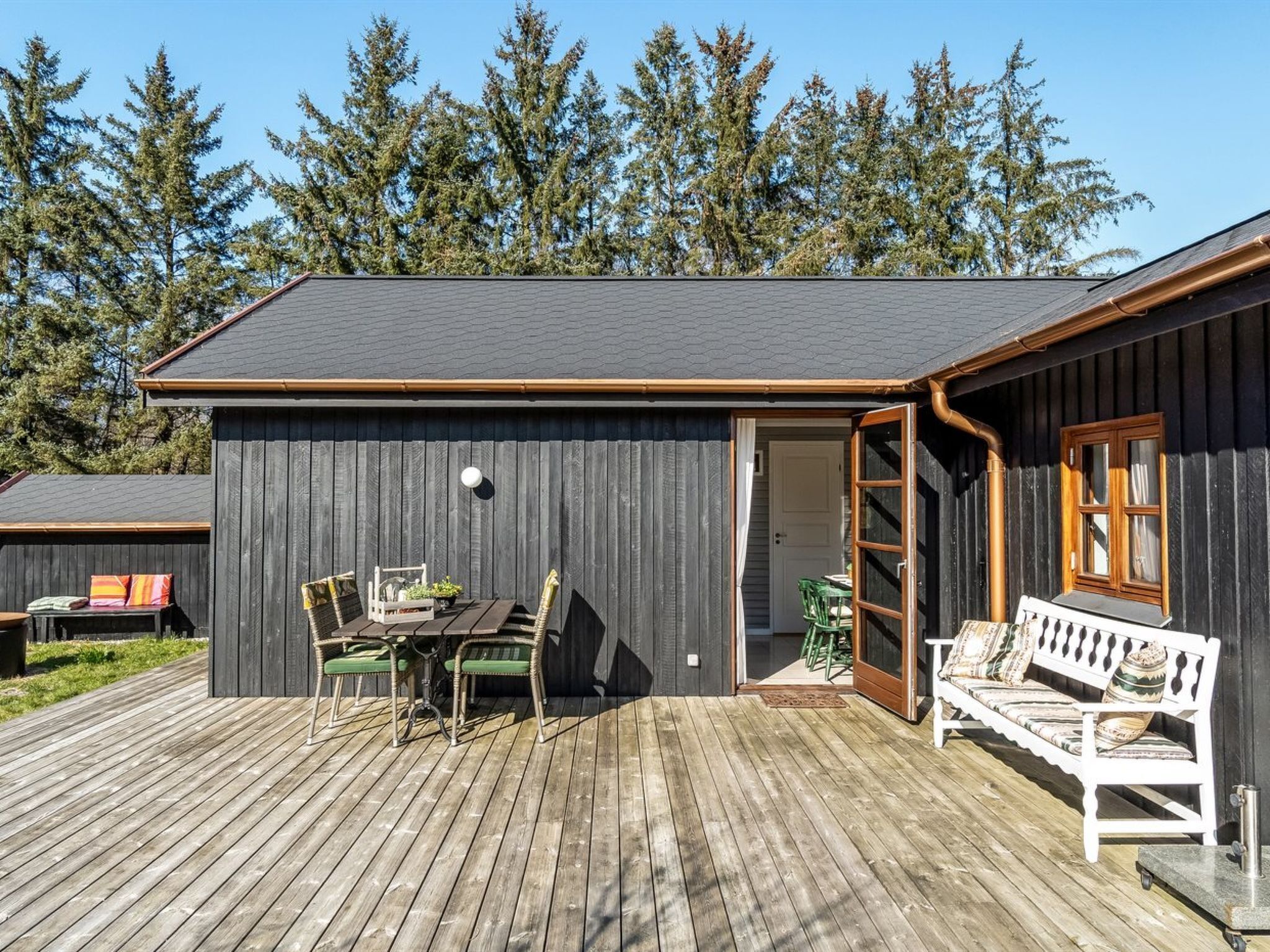 A wooden cabin with a deck, a dining area with chairs and a table, and a bench.