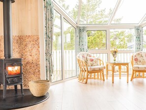A cozy room with a wood-burning stove, wicker basket, wicker chairs, and a view through large windows.