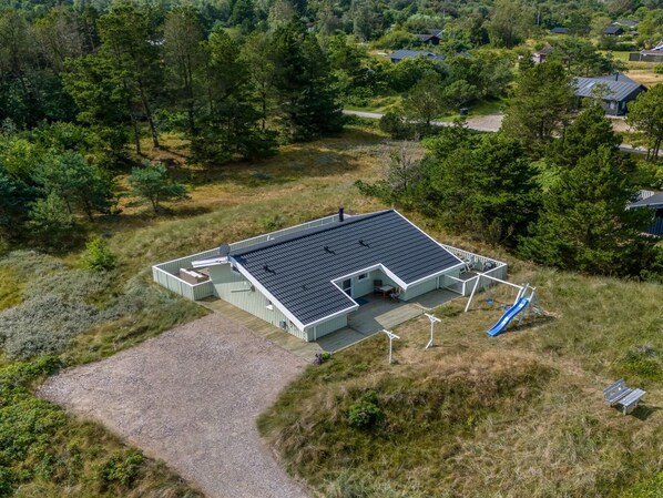 An aerial view of a modern, rectangular building with a flat roof, surrounded by dense greenery and a gravel driveway.