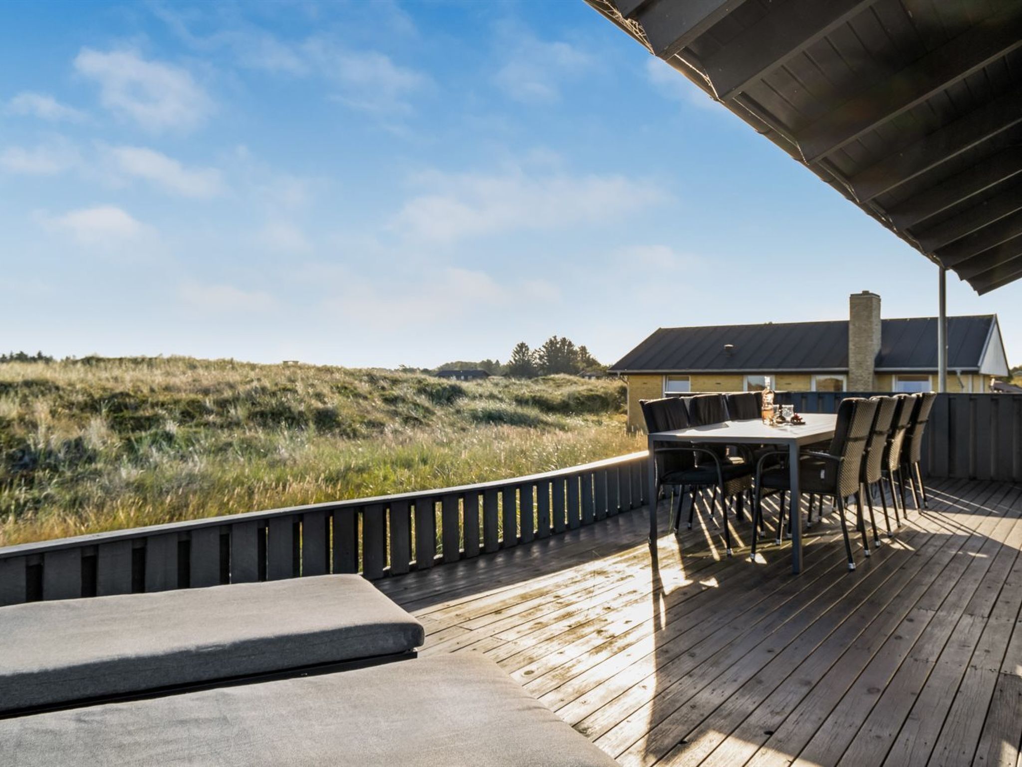 Una terraza de madera con una mesa y sillas, con vista a un paisaje cubierto de césped y un edificio.