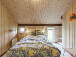 Un dormitorio con paredes y techo de madera, una cama con ropa de cama floral, una ventana y una silla.
