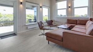 Un salon moderne avec un canapé en cuir marron, une petite table et un fauteuil. De grandes fenêtres laissent entrer la lumière naturelle et offrent une vue sur l’extérieur.
