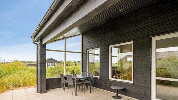 Une terrasse extérieure moderne, dotée d’une table et de chaises pour les repas, de grandes fenêtres et offrant une vue sur le paysage environnant.