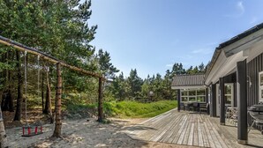 Een houten terras met buitenmeubilair, omringd door bomen en een zandgebied.
