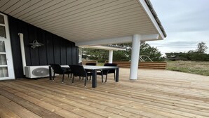Un espacio exterior cubierto con piso de madera, una mesa con sillas y una estructura de pilar blanco.