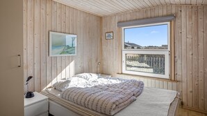 Ein Schlafzimmer mit Holzwänden, einem Bett mit Bettdecke, einem Nachttisch, einer Schreibtischlampe, einem gerahmten Bild und einem Fenster mit Blick auf einen Zaun und Gebäude.