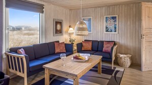 Ein gemütliches Wohnzimmer mit Holzbalkendecke, einer Sofagarnitur, einem Couchtisch und Blick durch ein großes Fenster.