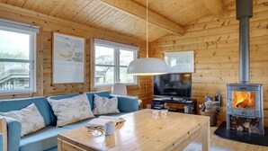 Ein gemütliches Blockhaus-Interieur mit Holzbalkendecke, einer Couch, einem Tisch, einem Fernseher und einem Kaminofen.