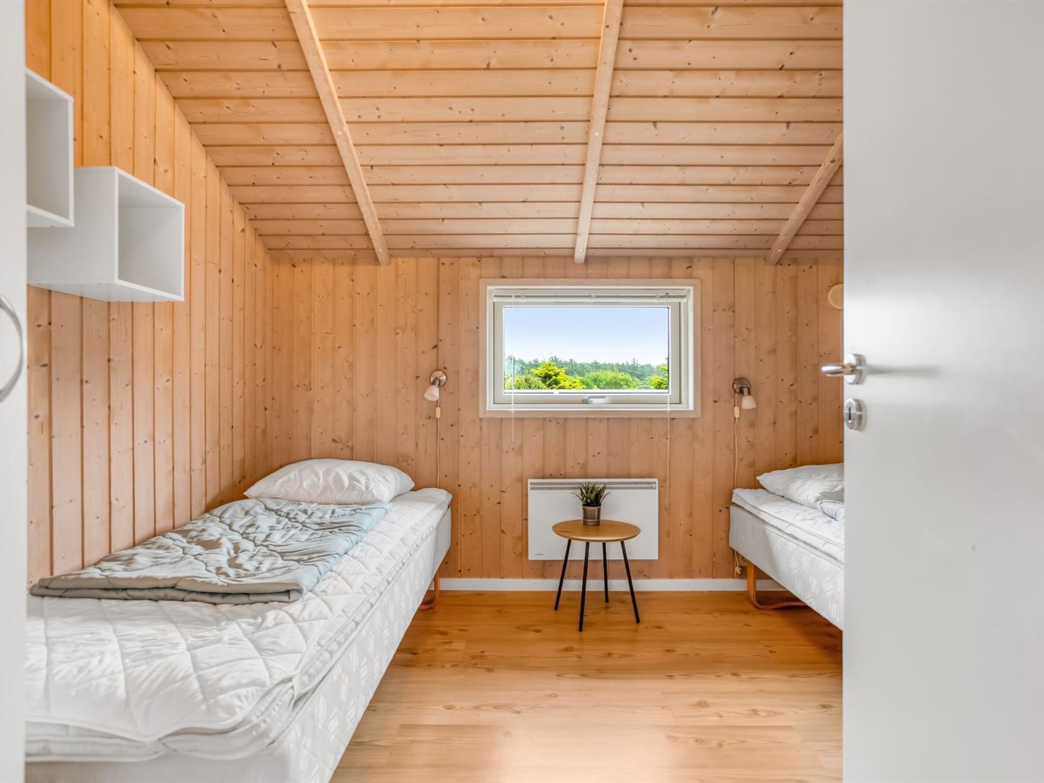 A small, wooden-paneled room with two beds, a window with a view of greenery, and a small table with a plant.