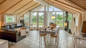 Een ruime woonkamer met hoge plafonds, houten balken, een leren bank, een eettafel met stoelen en een groot raam met uitzicht op het landschap.