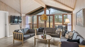 Ein geräumiges Wohnzimmer mit hoher Decke, Holzbalken, einem Flachbildfernseher, einem runden Couchtisch und einem großen Fenster mit Blick ins Freie.