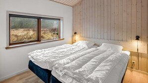 Ein Schlafzimmer mit Holzbalkendecke, einem großen Fenster mit Blick auf ein Feld, einem Bett mit weißer Bettwäsche und zwei Wandleuchten.