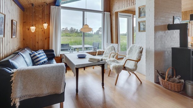 A cozy living room with a wooden ceiling, a fireplace, and a large window overlooking a grassy field.
