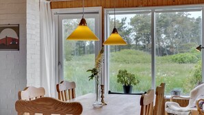 A dining area with a wooden table and chairs, two yellow pendant lights, a vase with flowers, and a view of greenery through large windows.