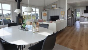 Ein geräumiger Wohnbereich mit einem weißen Esstisch, schwarzen Stühlen und einer zentralen Kochinsel. Im Hintergrund ist ein Kamin, ein Fernseher und eine Küche erkennbar.