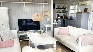 Ein Wohnzimmer mit einem weißen Sofa, einem weißen Couchtisch und einem Fernseher. Im Hintergrund ist eine Küche mit weißen Schränken und einem Fenster zu sehen.