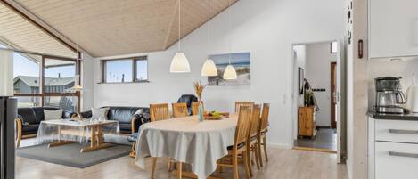 Een ruime woonkamer met een eettafel, houten stoelen, een bank en een keukenhoek met koffiezetapparaat.