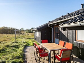 Una casa de madera con techo de tejas, un patio con mesa y sillas, y un campo cubierto de césped al fondo.