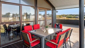 Un espace de restauration extérieur abrité, avec une table en bois et des chaises rouges, de grandes fenêtres et une vue sur le paysage environnant.