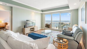 Un salon moderne avec un canapé, un pouf et une table basse en verre. On y trouve un téléviseur posé sur un meuble en bois, ainsi qu’une grande fenêtre donnant sur la mer.
