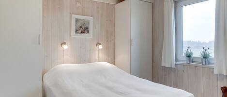 Ein Schlafzimmer mit einem Bett, zwei Nachttischlampen, einem Fenster mit Vorhängen und einem gerahmten Bild an der Wand.