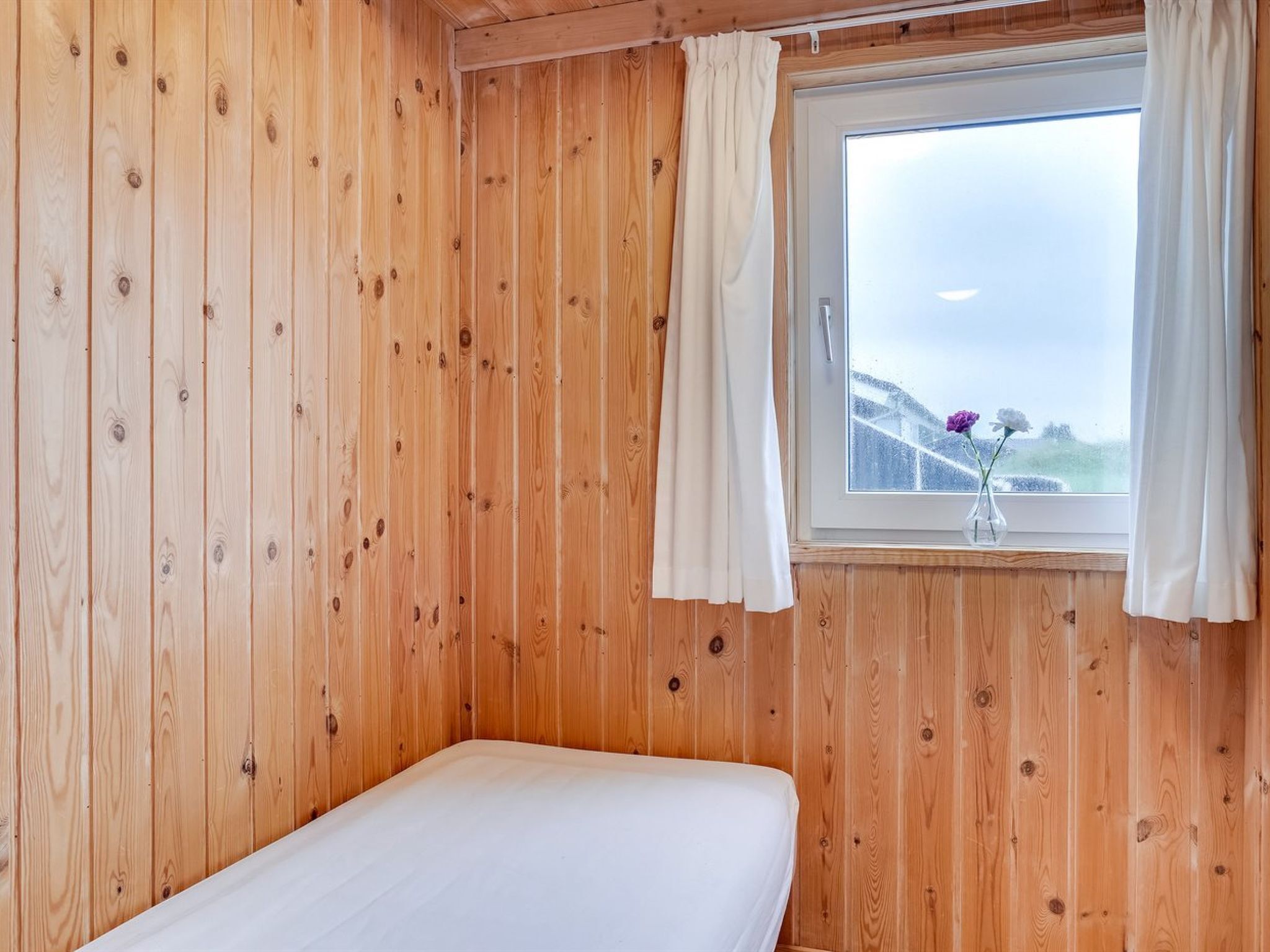 A room with wooden walls, a single bed, a window with curtains, and a view of a snowy landscape.
